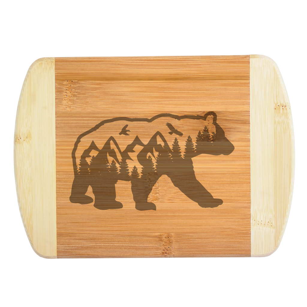 A premium engraved bamboo cutting board featuring an intricate illustration of a grizzly bear silhouette filled with mountains, evergreen trees, and flying eagles, handcrafted by The Blended Oasis in Washington State.