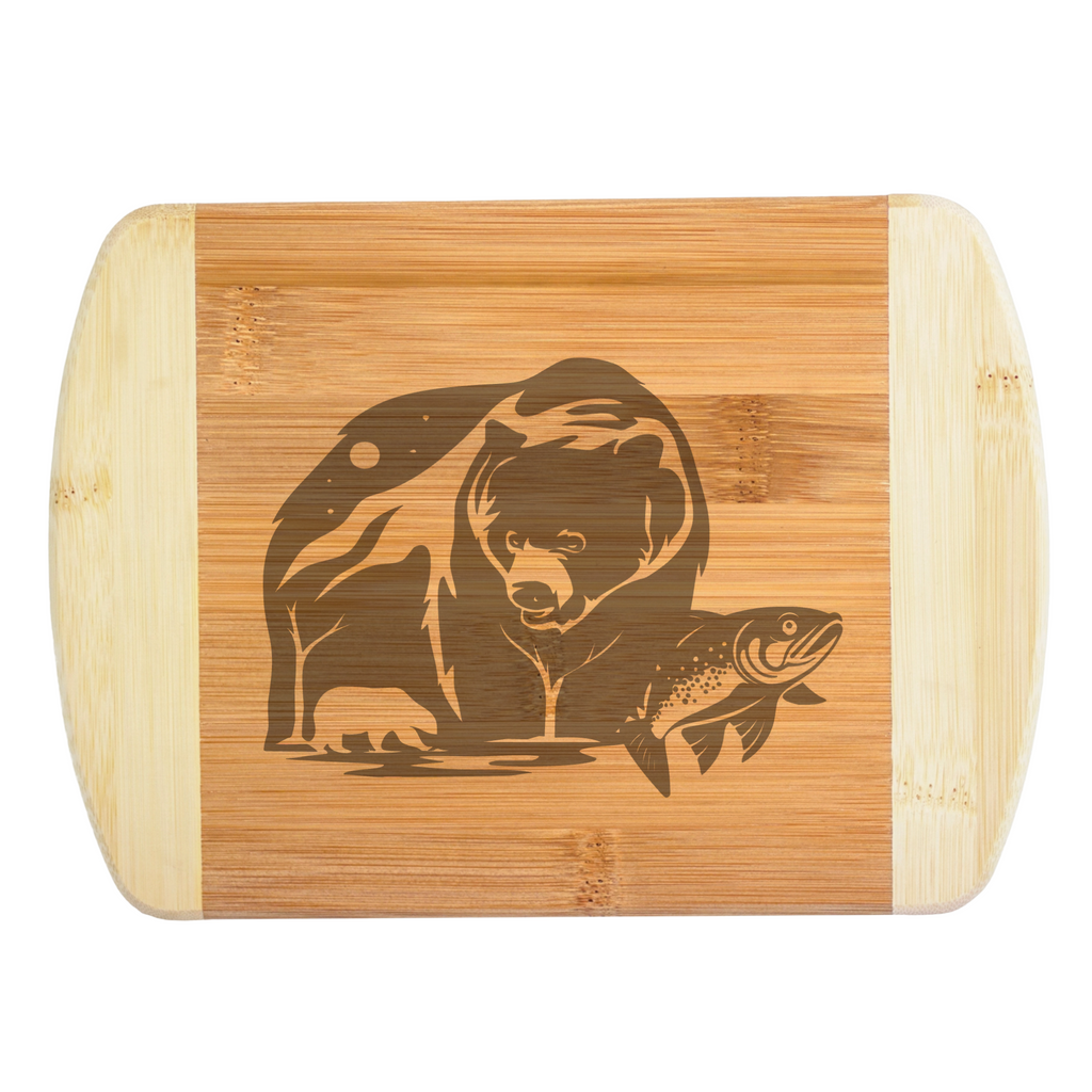 A premium engraved bamboo cutting board featuring a detailed illustration of a grizzly bear, a small cub, and a leaping salmon fish by a riverbank, handcrafted by The Blended Oasis in Washington State.