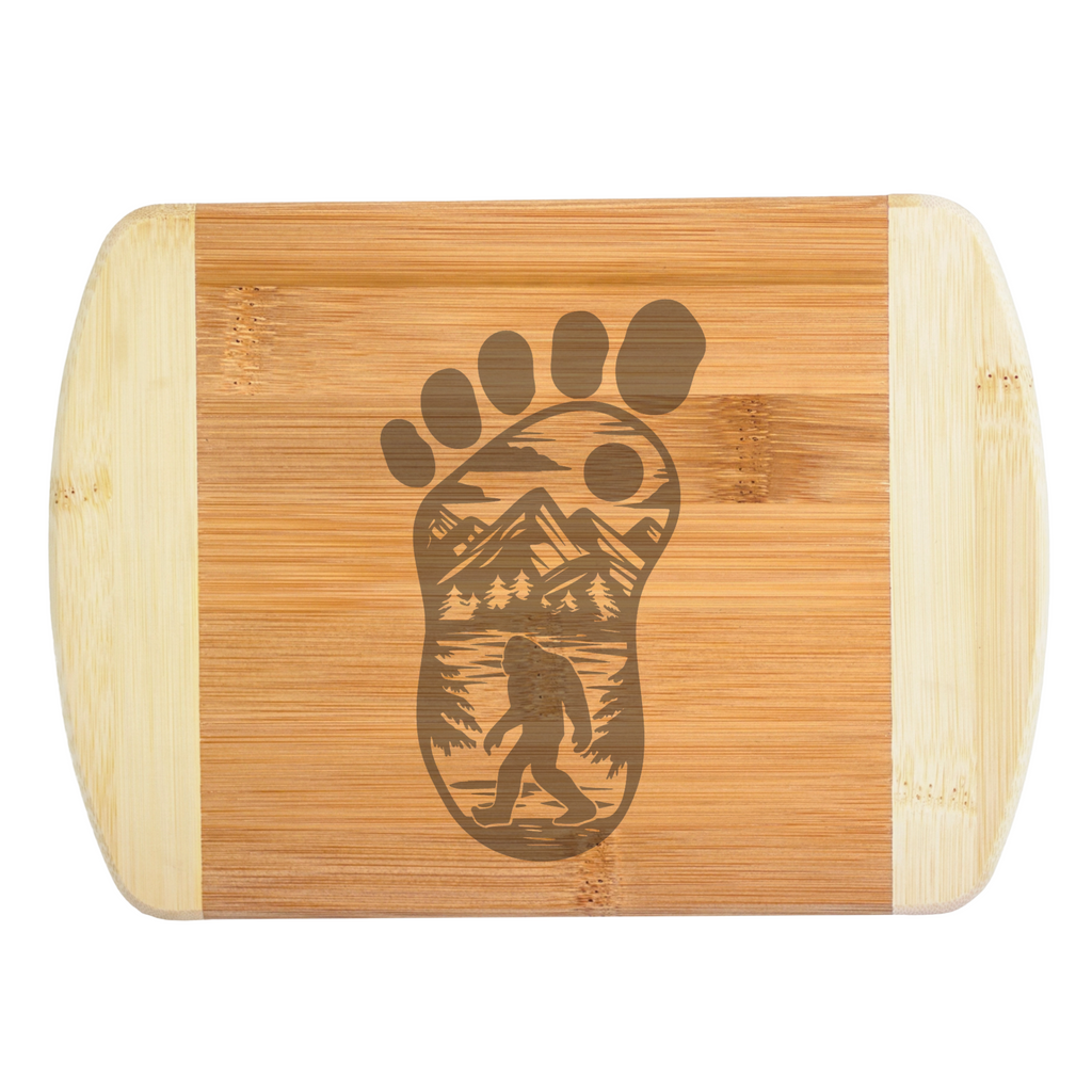 A premium engraved bamboo cutting board featuring an intricate illustration of a giant footprint filled with mountains, trees, and a walking Bigfoot silhouette, handcrafted by The Blended Oasis in Washington State.