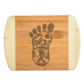 A premium engraved bamboo cutting board featuring an intricate illustration of a giant footprint filled with mountains, trees, and a walking Bigfoot silhouette, handcrafted by The Blended Oasis in Washington State.