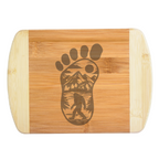 A premium engraved bamboo cutting board featuring an intricate illustration of a giant footprint filled with mountains, trees, and a walking Bigfoot silhouette, handcrafted by The Blended Oasis in Washington State.