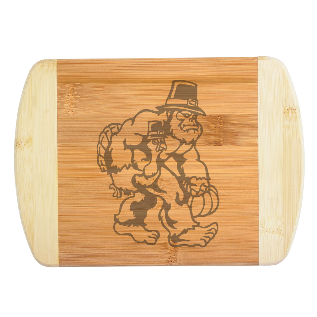 A premium engraved bamboo cutting board featuring a funny autumn illustration of Bigfoot wearing a buckled hat while carrying a turkey and a pumpkin, handcrafted by The Blended Oasis in Washington State.