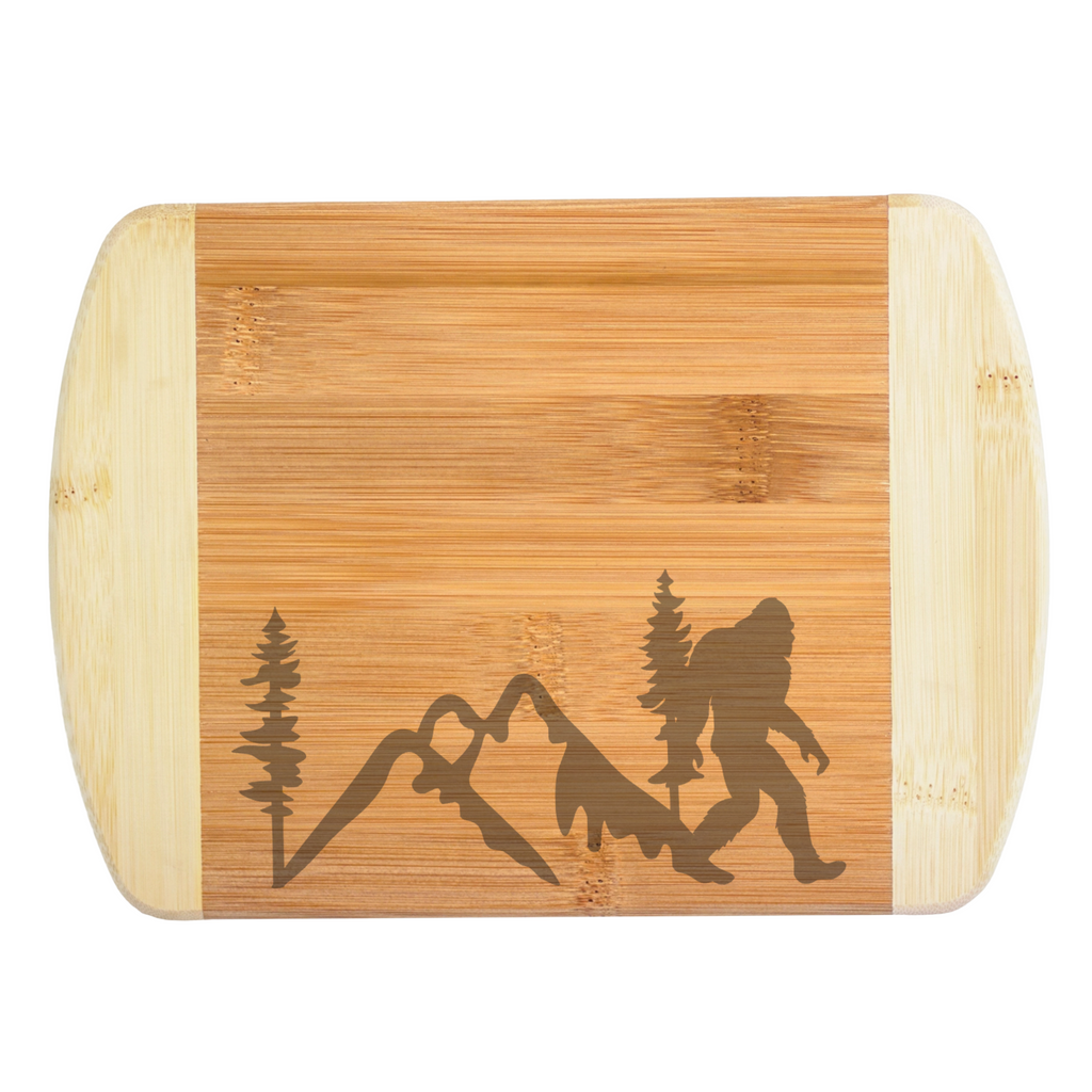 A premium engraved bamboo cutting board featuring a clean, low-profile baseline illustration of mountain peaks, pine trees, and a walking Bigfoot silhouette, handcrafted by The Blended Oasis in Washington State.