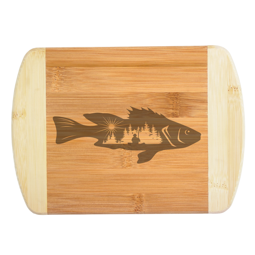 A rustic laser-engraved bamboo cutting board featuring a unique hybrid design of a fish silhouette filled with a detailed lakeside wilderness scene, a fisherman in a kayak, and a rising sun, handcrafted by The Blended Oasis in Washington State.