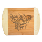 A beautiful engraved bamboo cutting board styled on a kitchen counter featuring a unique split honeybee design with a dense trail of wildflowers forming half its body, handcrafted by The Blended Oasis in Washington State.