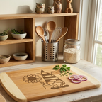 A handcrafted bamboo cutting board featuring a laser-engraved gnome holding a large sunflower, displayed in a rustic kitchen setting. The board is two-toned with light bamboo end caps and a darker center, part of a functional prep scene with sliced red onion, chopped parsley, a chef's knife, open wooden shelves with fresh herb bowls, and natural light from a window.
