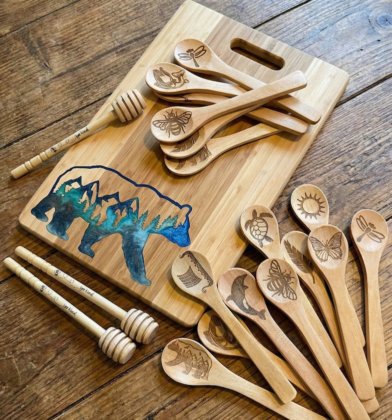 A handcrafted bamboo cutting board featuring a blue epoxy resin bear inlay with a mountain forest scene, surrounded by wooden honey dippers engraved with 'bee kind' and laser-engraved wooden spoons showing nature designs like bees and dragonflies, created by The Blended Oasis in Suquamish, Washington.