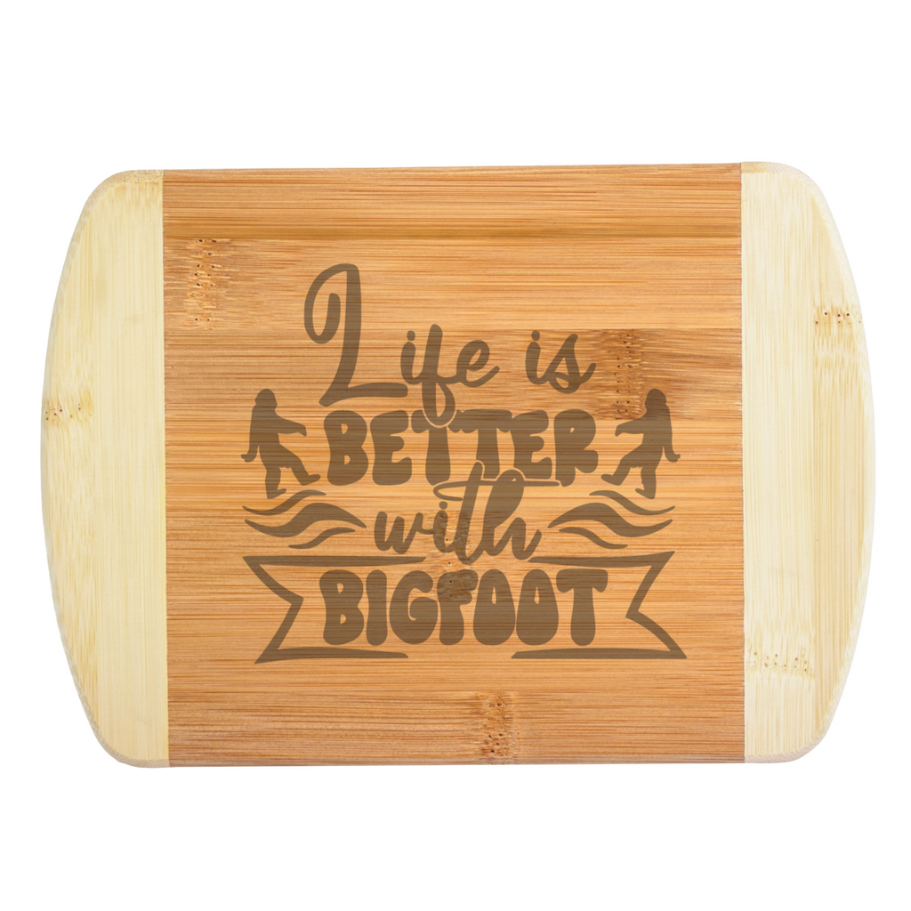 A premium engraved bamboo cutting board featuring a playful retro typography layout that reads Life is Better with Bigfoot, flanked by two small Sasquatch silhouettes, handcrafted by The Blended Oasis in Washington State.