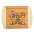 A premium engraved bamboo cutting board featuring a playful retro typography layout that reads Life is Better with Bigfoot, flanked by two small Sasquatch silhouettes, handcrafted by The Blended Oasis in Washington State.