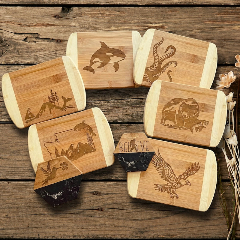 A collection of custom laser-engraved bamboo cutting boards by The Blended Oasis featuring Pacific Northwest wildlife, including an orca, octopus, bear, eagle, and a Washington state outline. The display also includes two hexagonal wood and black epoxy resin coasters showing a hummingbird and a Sasquatch silhouette with the word BELIEVE, all arranged on a rustic wooden table.
