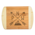 A premium engraved bamboo cutting board featuring a trendy modern illustration of crossed arrows with the letters PNW and a clean mountain peak silhouette, handcrafted by The Blended Oasis in Washington State.