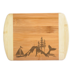 A premium engraved bamboo cutting board featuring a unique baseline illustration collage of a sailboat, mountain peak, evergreen pine trees, and a whale tail fluke, handcrafted by The Blended Oasis in Washington State.