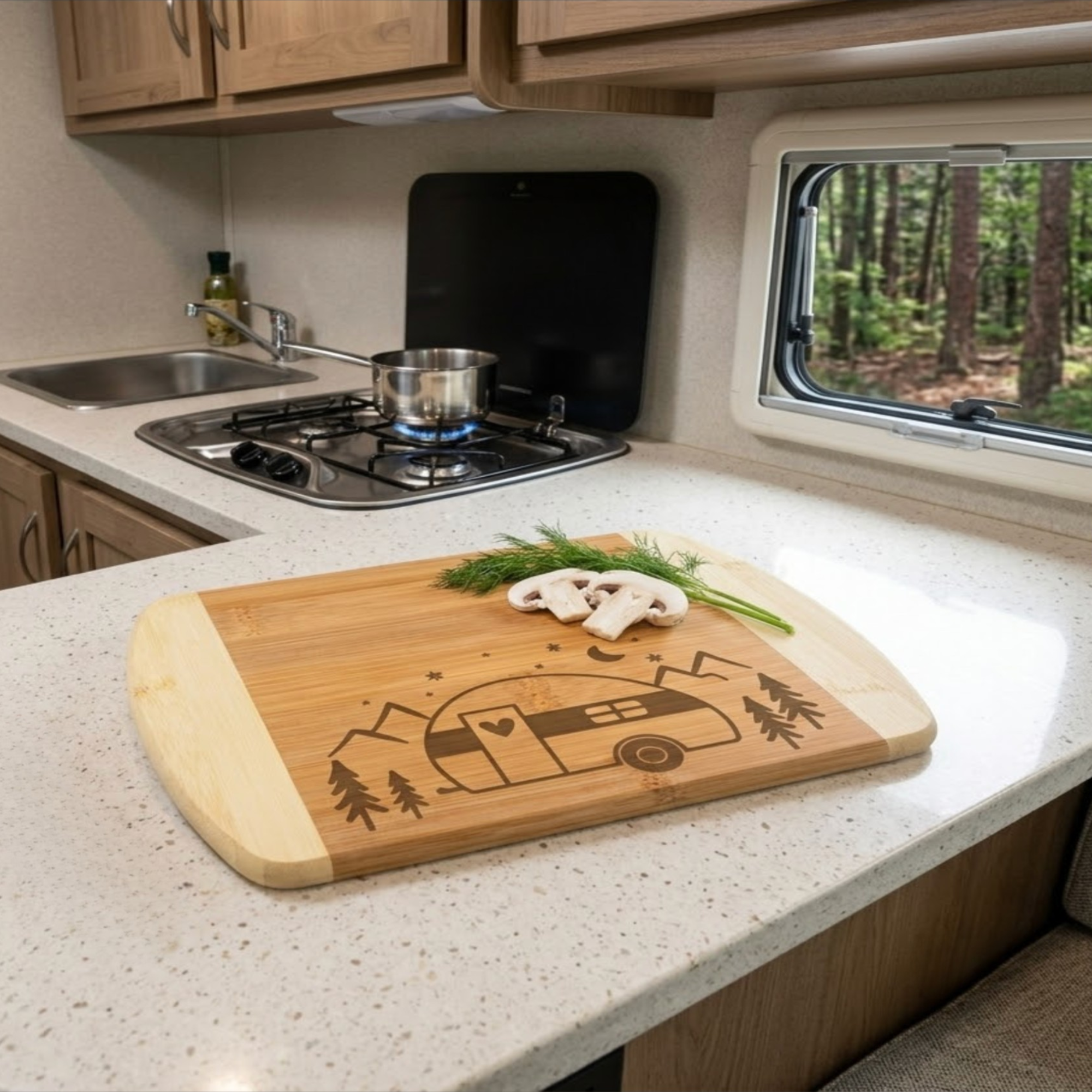 Wooden cutting board with camping design on a kitchen counter with stove and window view. Two-tone bamboo cutting board laser-engraved with a retro camper, mountains, and trees, displayed on a light countertop inside a compact RV kitchen by The Blended Oasis.