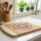 A two-tone bamboo cutting board featuring a large laser-engraved sunflower with a mountain and evergreen tree scene in the center, displayed on a white kitchen counter next to fresh vine tomatoes and wooden utensils by The Blended Oasis