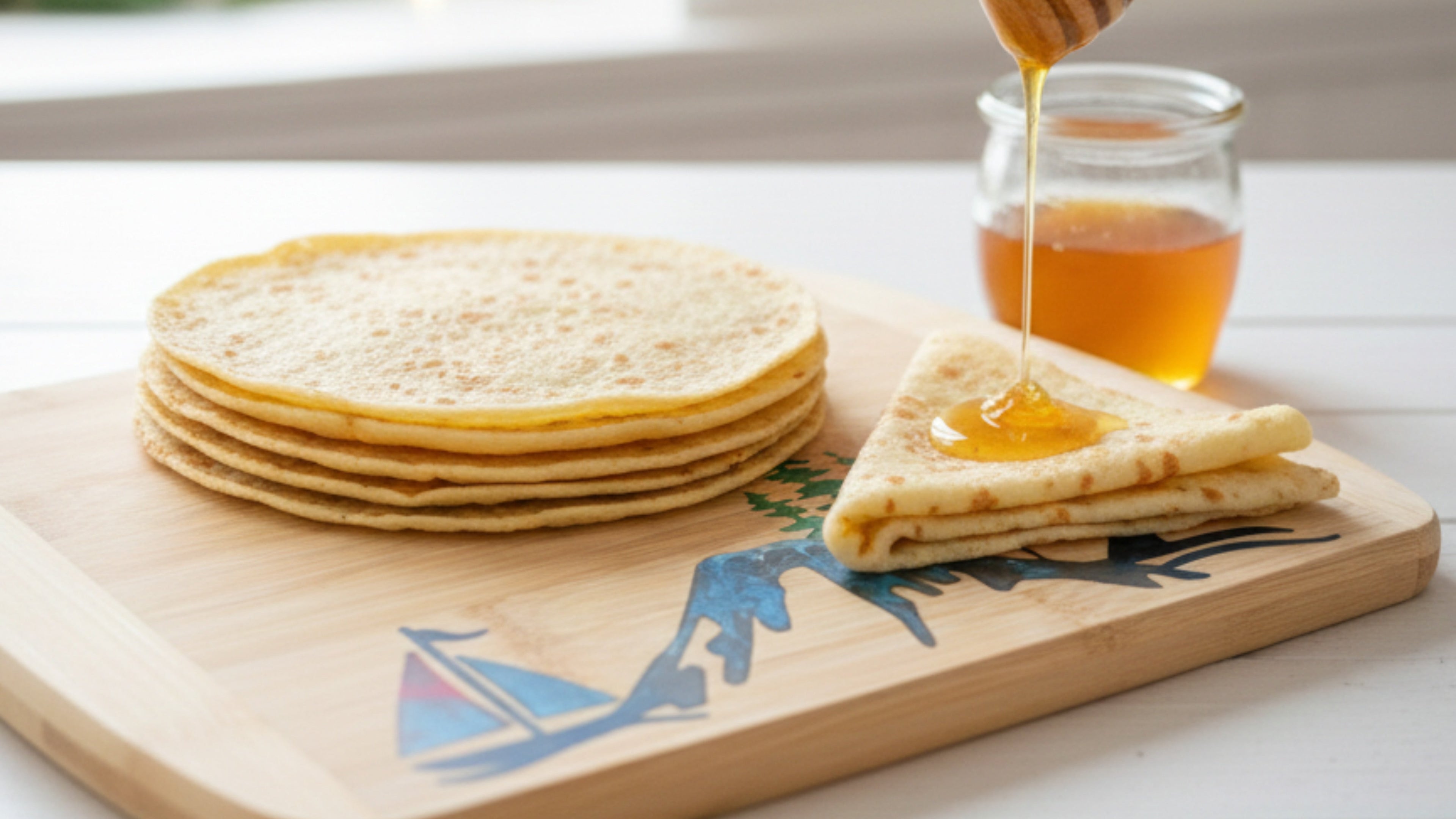 A close-up of a bamboo cutting board from The Blended Oasis, featuring a subtle mountain and boat design, holding a stack of crepes. Honey is being drizzled onto a folded crepe with a honey dipper, next to a jar of honey. A soft, festive background with blurry lights creates a cozy, inviting kitchen scene.