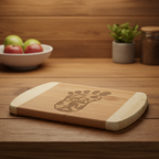 A bamboo cutting board by The Blended Oasis featuring a clever laser engraving of a large Bigfoot footprint, with a detailed silhouette of Sasquatch walking through a forest etched inside it. Shown on a warm, dark wood counter next to a bowl of apples.
