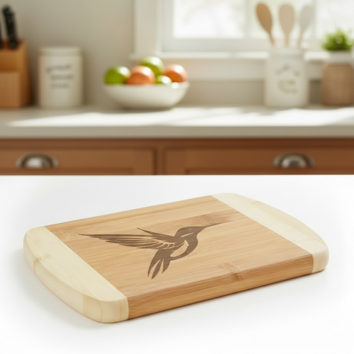 A bright, high-key product photo of a bamboo cutting board by The Blended Oasis, featuring a detailed laser etching of a hummingbird in flight. Shown on a crisp white surface with a soft kitchen background.