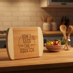 A humorous bamboo serving board from The Blended Oasis featuring bold typographic laser engraving that reads "Don't Sass the Squatch" inside a graphic circular frame. The board stands upright on a warm wood countertop next to a jar of wooden cooking spoons.