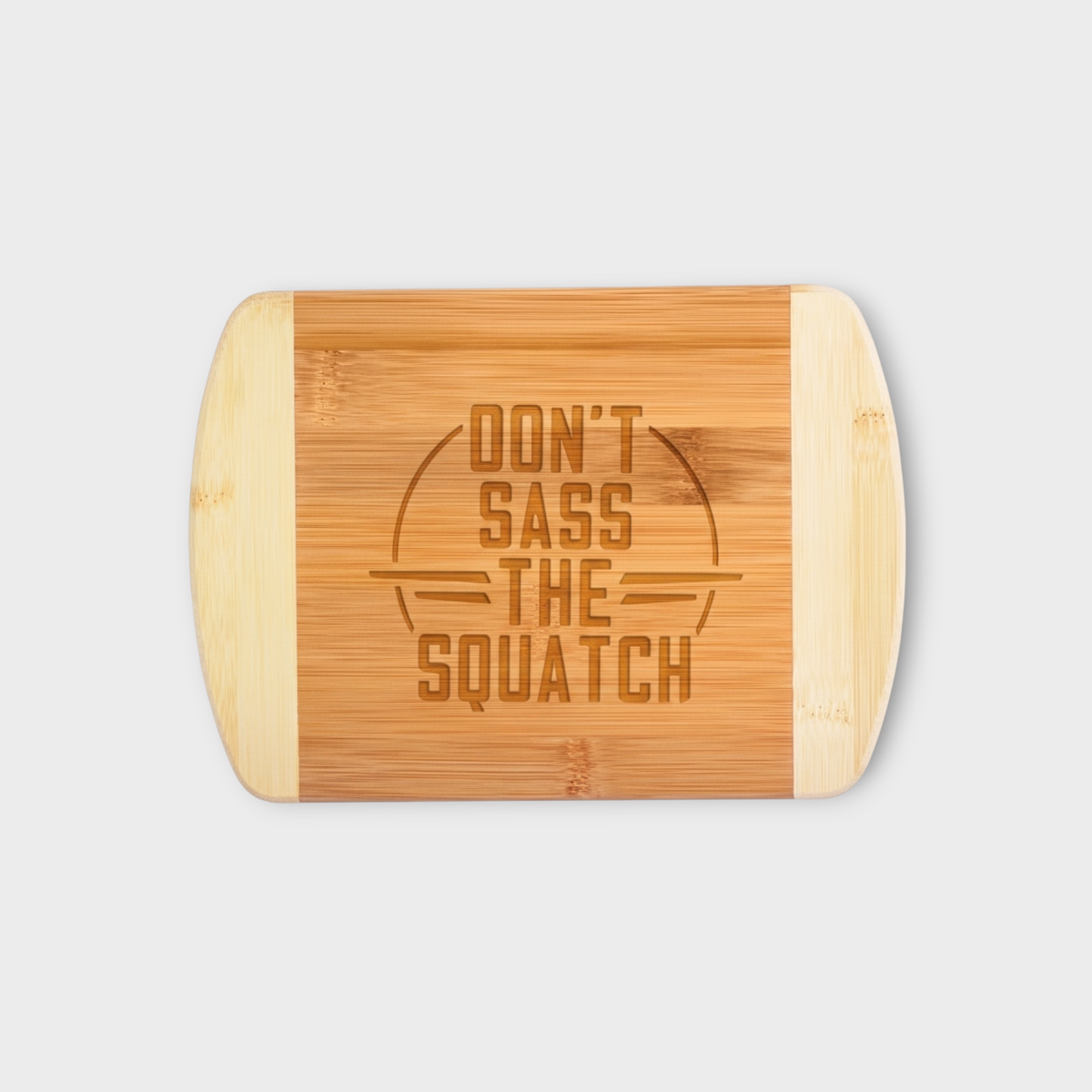 A rectangular bamboo cutting board with rounded corners, featuring a natural wood center panel engraved with the phrase 'DON'T SASS THE SQUATCH' inside a circle, flanked by two lighter bamboo end panels, shown on a light gray background.