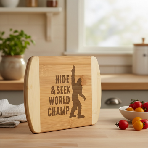 A humorous bamboo cutting board by The Blended Oasis, laser-engraved with a silhouette of Sasquatch throwing a peace sign next to the text "Hide & Seek World Champ." The board stands upright on a kitchen table next to small tomatoes.