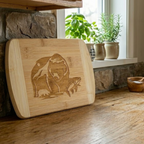 A rustic bamboo cutting board from The Blended Oasis featuring a detailed laser engraving of a grizzly bear with a salmon in its mouth, set against a mountain backdrop. The board leans against a stone fireplace surround near a sunlit window with potted herbs.