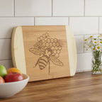  An artisanal wooden charcuterie board from The Blended Oasis, showcasing a detailed laser-etched bee on a honeycomb pattern. The board leans against a white subway tile backsplash next to a bowl of fresh fruit.
