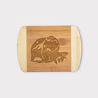A rectangular bamboo cutting board with rounded corners and handles, featuring a laser-engraved graphic of a bear and a fish in the center, shown against a light gray background.