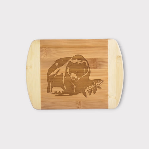 A rectangular bamboo cutting board with rounded corners and handles, featuring a laser-engraved graphic of a bear and a fish in the center, shown against a light gray background.