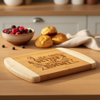 A custom cutting board by The Blended Oasis with a beautiful typographic laser engraving that reads "Life is Better with Bigfoot" surrounded by wilderness details. Displayed on a table next to a bowl of fresh berries and warm biscuits.