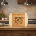 A nautical-themed bamboo cutting board from The Blended Oasis featuring a striking laser-etched ship's steering wheel intertwined with delicate floral blooms. The board stands upright on a rustic wooden bar top against a soft kitchen backdrop.