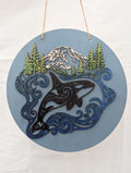 Decorative wall hanging with an orca whale, mountains, and trees on a blue background.