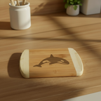 A premium laser-engraved serving board from The Blended Oasis, featuring a sleek Pacific Northwest killer whale design. The bamboo board rests on a sunlit wooden counter with sharp geometric shadows casting across the surface.