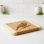 A handmade wooden cutting board by The Blended Oasis, featuring a laser-engraved Pacific Northwest songbird perched on a branch. The board is shown at an angle on a clean, bright white kitchen countertop.