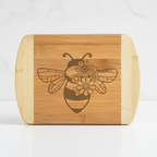 A straight-on product shot of a personalized bamboo serving board by The Blended Oasis, featuring a bold, graphic laser-engraved bee with floral wings centered between light wood accents.