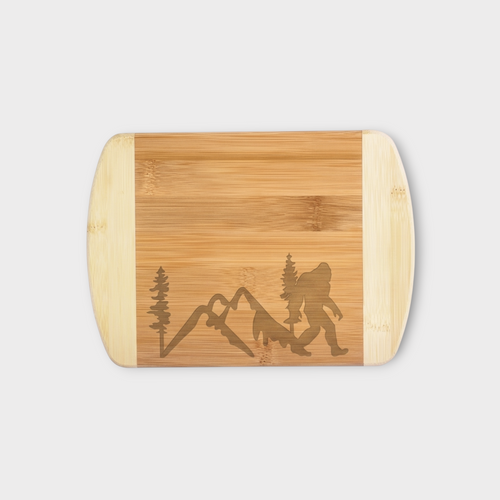 A rectangular bamboo cutting board with rounded corners, featuring a natural wood grain surface with an engraved silhouette of a hiking backpacker, mountain peaks, and pine trees. The ends of the board are made of a lighter, pale wood tone.