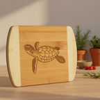 A beautiful product photo of a bamboo serving board from The Blended Oasis, featuring a detailed laser-etched sea turtle design. The board stands upright on a sunlit table with small potted rosemary plants in the soft background.