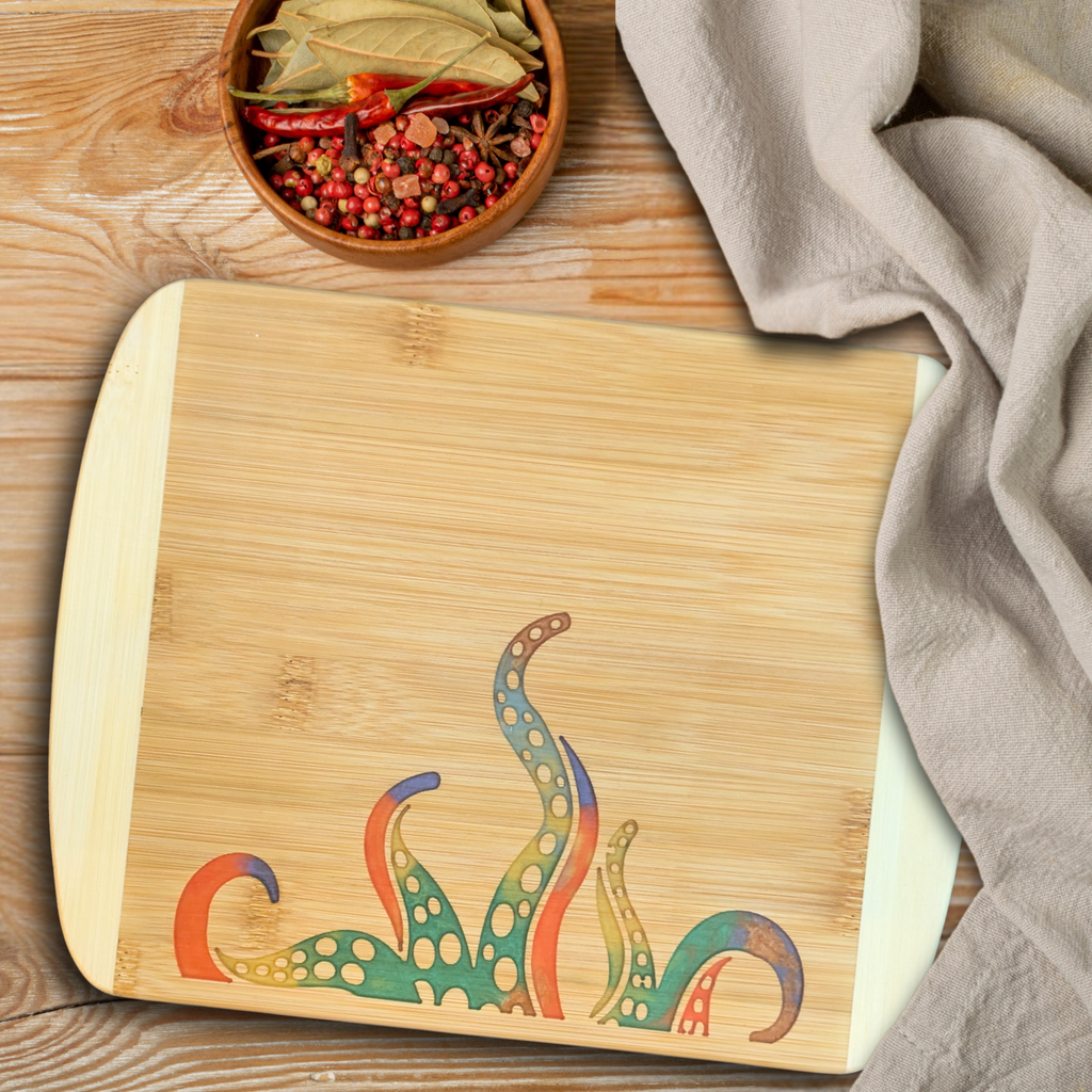 Handcrafted bamboo cutting and serving board by The Blended Oasis in Suquamish, WA, featuring a vibrant, multi-colored epoxy resin inlay of stylized kraken tentacles rising from the bottom edge. Styled on a light wood surface next to a neutral cloth napkin and a small wooden bowl filled with dried chili peppers and peppercorns.