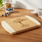 A beautiful bamboo serving board by The Blended Oasis featuring a detailed laser engraving of a mystical Tree of Life where the trunk forms a woman's silhouette with deep roots. Sits on a warm wooden countertop next to a bowl of fresh blueberries.