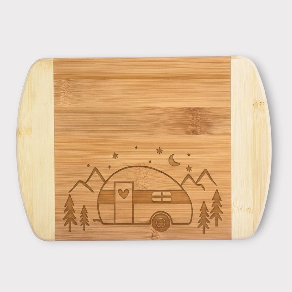 A rectangular bamboo cutting board with rounded corners and handles, featuring a laser-engraved design of a vintage camper trailer, mountains, stars, and crescent moon on the front, set against a light gray background.