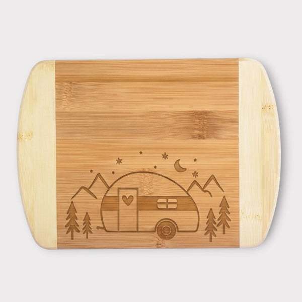 A rectangular bamboo cutting board with rounded corners and handles, featuring a laser-engraved design of a vintage camper trailer, mountains, stars, and crescent moon on the front, set against a light gray background.