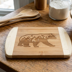 A premium bamboo cutting board from The Blended Oasis featuring a laser-engraved silhouette of a walking bear filled with a majestic mountain range and pine forest. The board rests flat on a wooden kitchen counter next to large wooden spoons and a jar of flour.