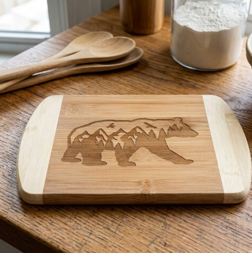 A premium bamboo cutting board from The Blended Oasis featuring a laser-engraved silhouette of a walking bear filled with a majestic mountain range and pine forest. The board rests flat on a wooden kitchen counter next to large wooden spoons and a jar of flour.